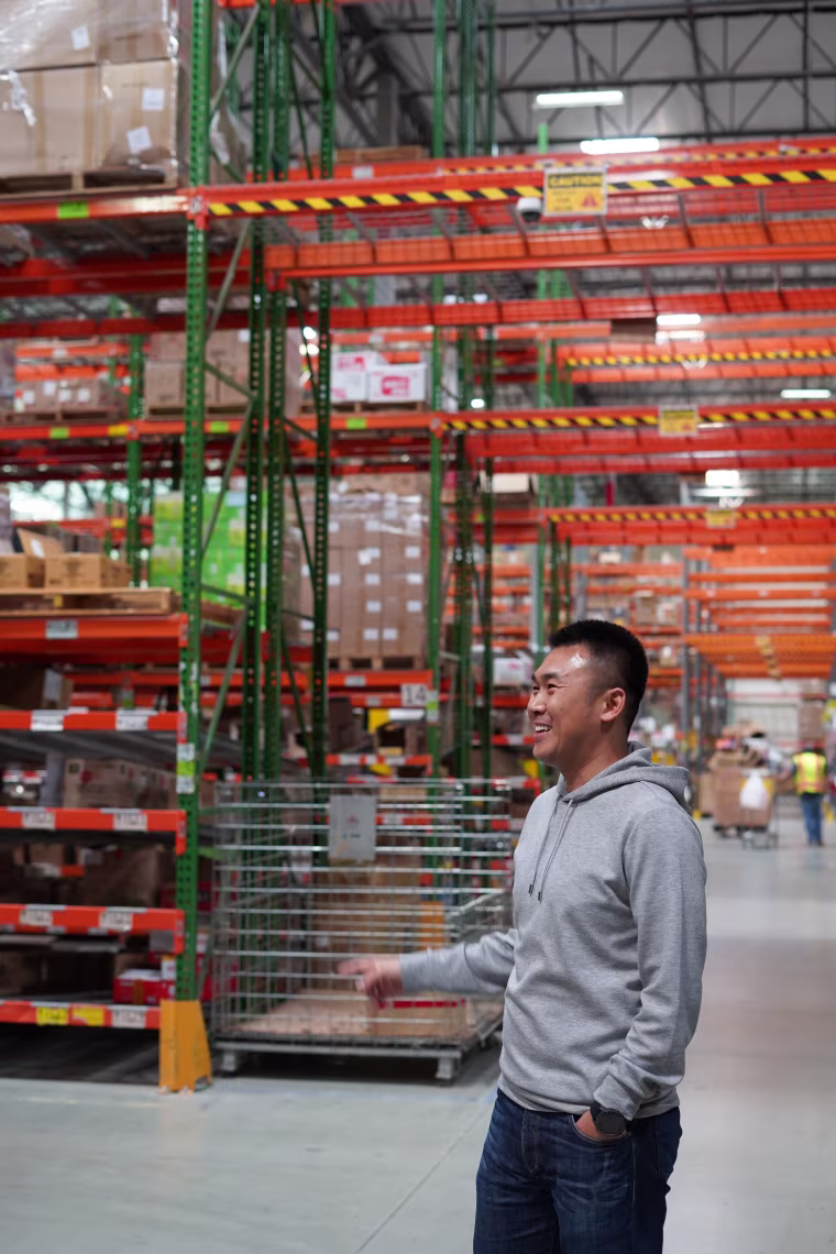 A young Asian man stands in front of shelves in a warehouse.