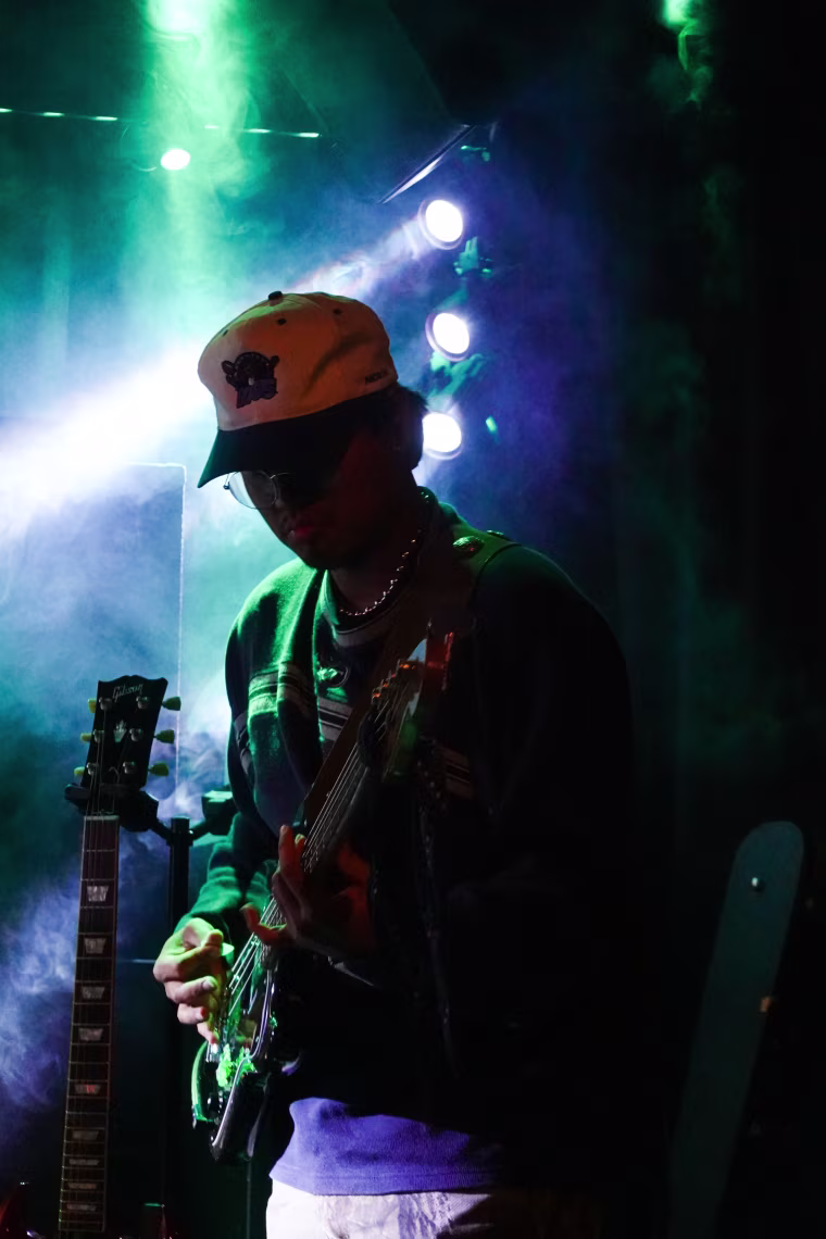 A photo of a musician playing electric guitar on a stage with blue and green flood lights in the background.