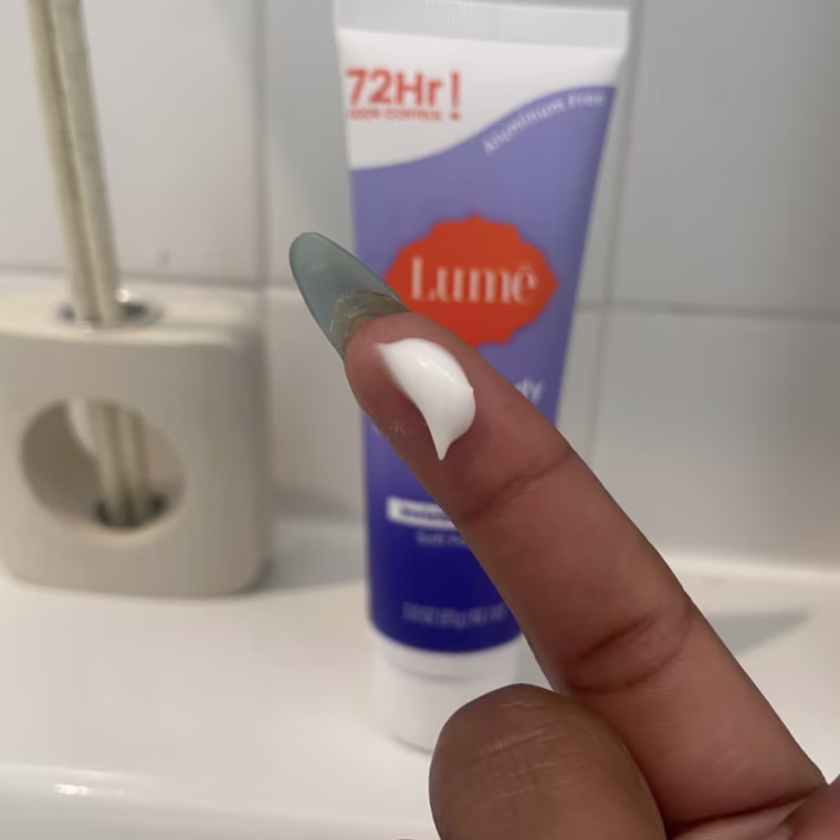 A drop of the Lume Whole Body Deodorant shown on a fingertip in front of a sink, with the deodorant tube on the counter in the background.