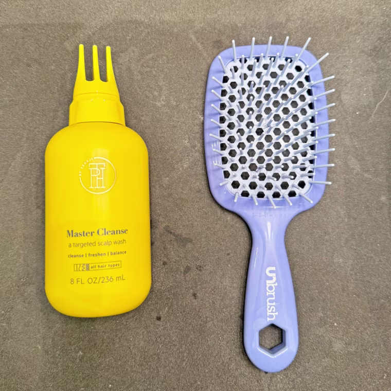 On the left is the yellow TPH By Taraji Master Scalp Shampoo bottle, and on the right is a light purple vented paddle brush from FHI Heat.