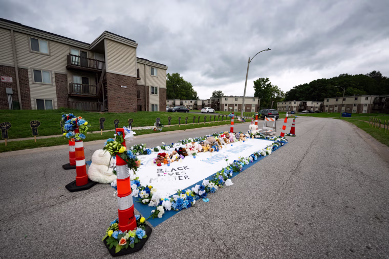 A memorial for Michael Brown in the center of a street near apartment building is laden with flowers, cones, and text that reads "Black Lives Matter" and "Mike Brown"
