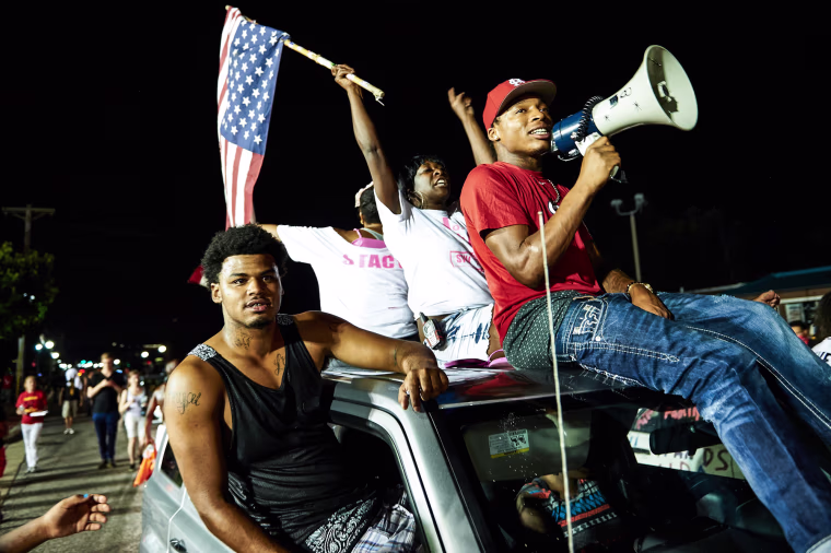 Demonstrators protest on the street, a woman waves an American flag sitting atop a car next to a man holding a megaphone