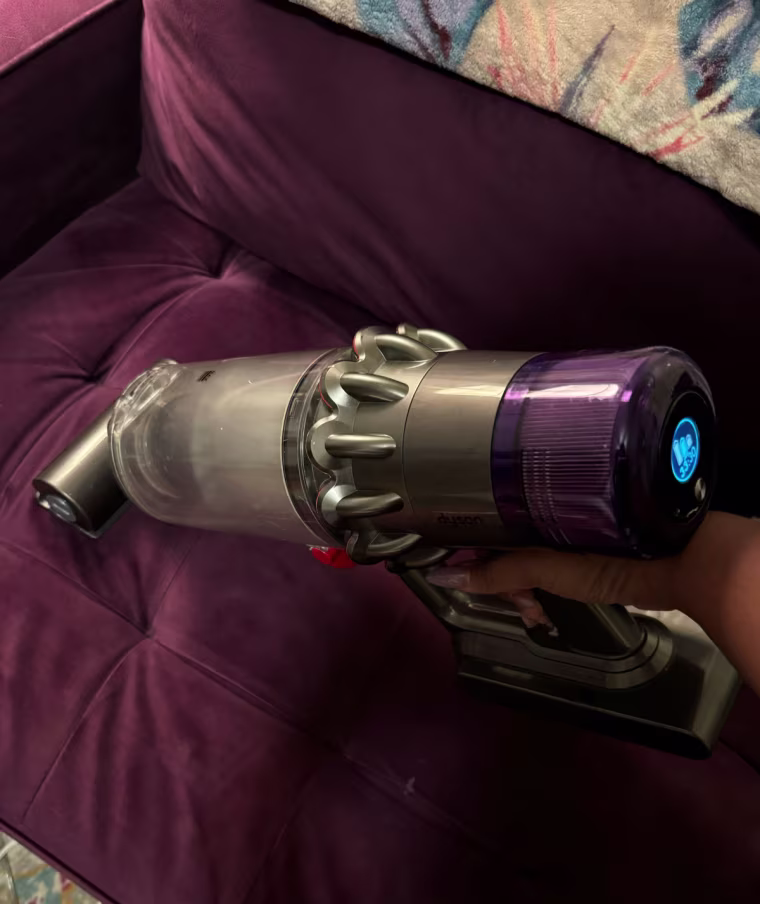 A close up of a Dyson vacuum being used on a purple couch in handheld mode.