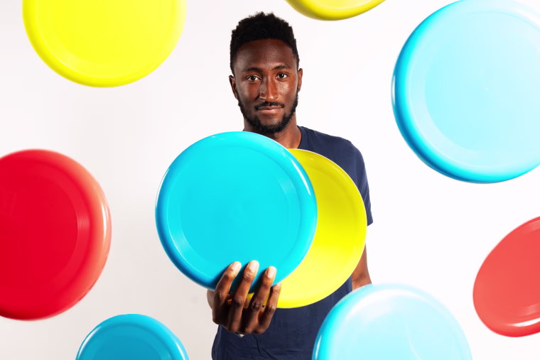 Marques Brownlee holding two frisbees with multicolored frisbees in the air around him.