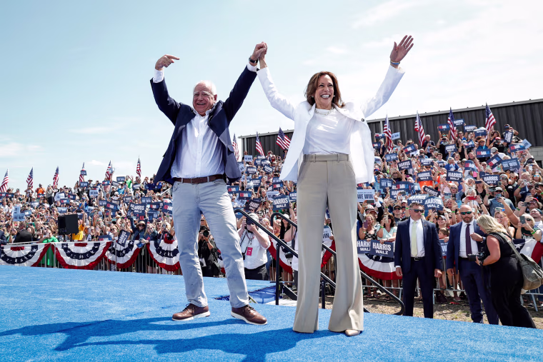 TIm Walz and Kamala Harris on stage outside, hold hands and raise their arms in celebration