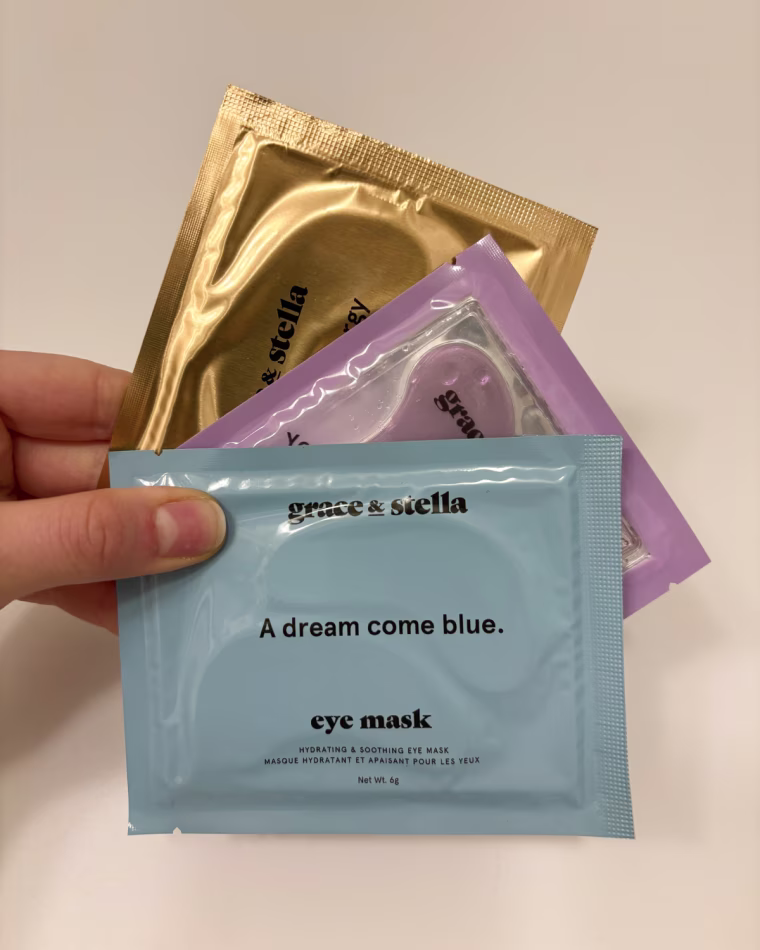 A woman’s hand holding three packages of Grace & Stella under-eye patches against a white wall: Energizing (top), Restoring (middle) and Illuminating (bottom).