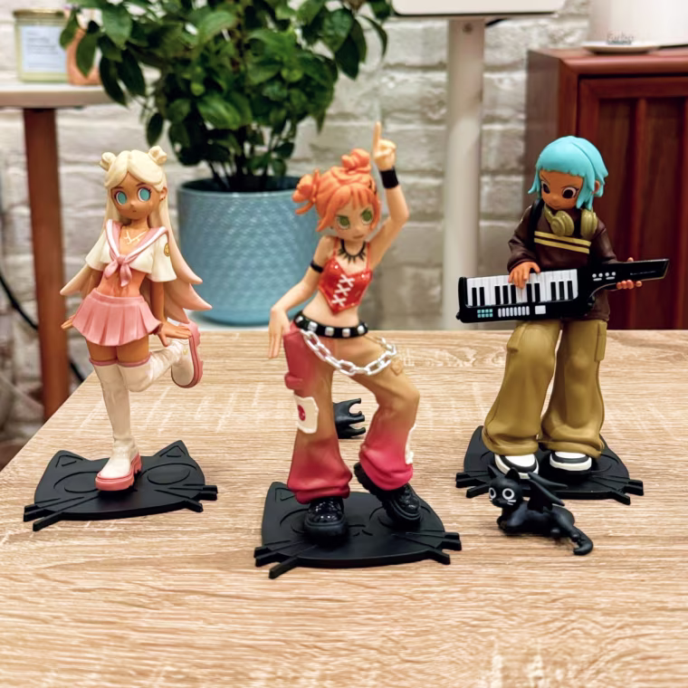 Three Peach Riot Rise Up figurines on a table inf front of a brick wall, a house plant and a speaker.