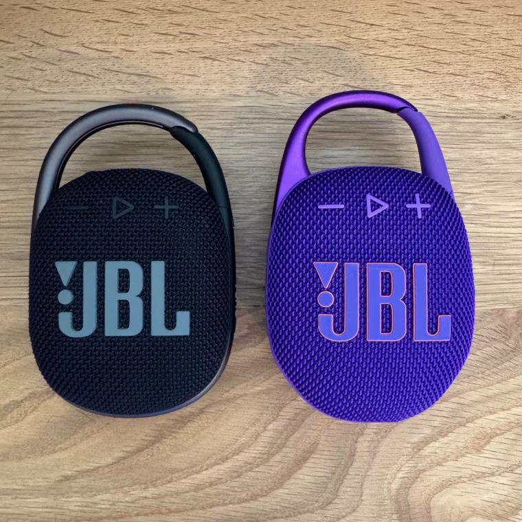 Purple oval-shaped JBL speaker with carabiner clip on the right and slightly smaller black oval-shaped JBL speaker with carabiner clip on the left.