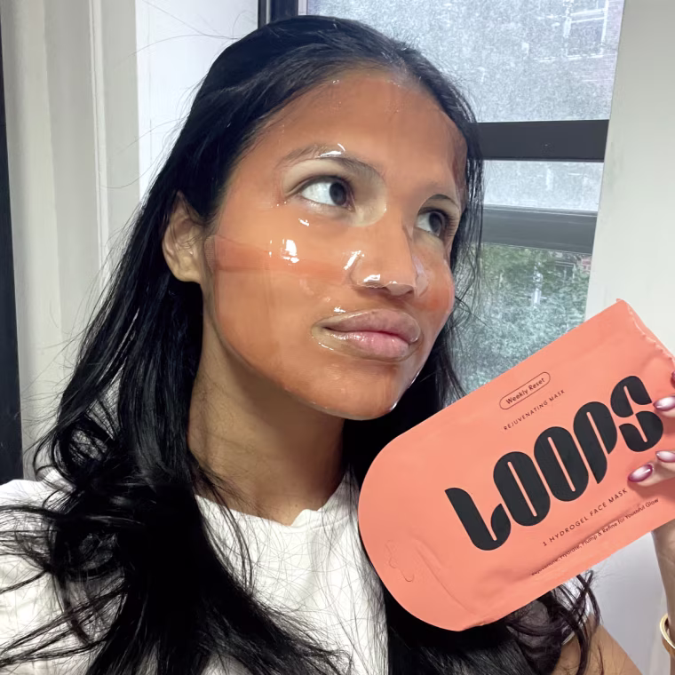 A woman wearing a Loops sheet mask while holding up it's packaging in her hand.