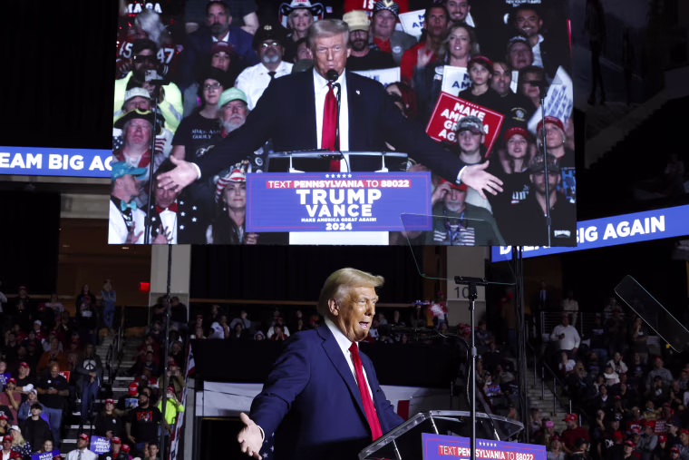 Donald Trump speaks in front of a big screen showing him.
