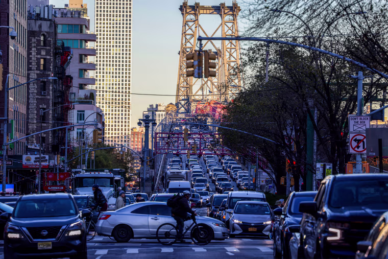 Pedestrians, cyclists and cars move along Delancey Street with the Williamsburg Bridge in the background,