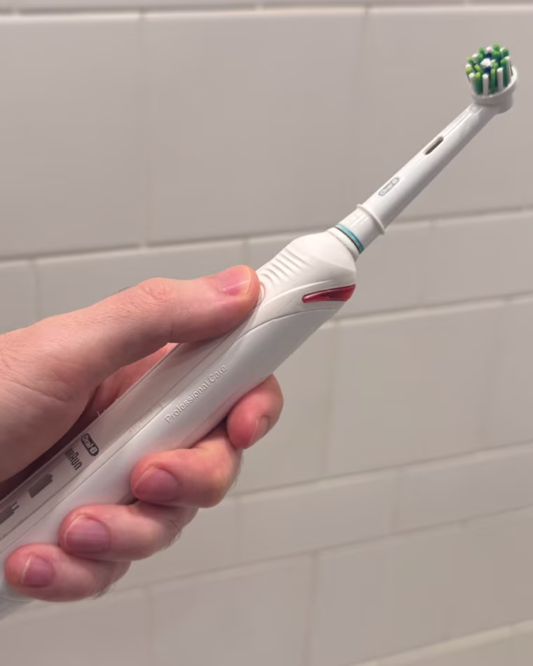 A man’s hand holding the Oral-B Smart 1500 Electric Toothbrush against a white tile wall.