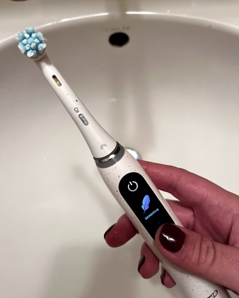 A woman’s hand holding the Oral-B iO Series 10 Electric Toothbrush over a bathroom sink with the word “sensitive” displayed on the handle’s screen.