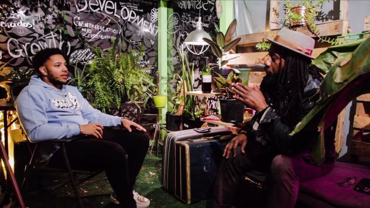 Two men speak to each other while seated inside of a room with various plants