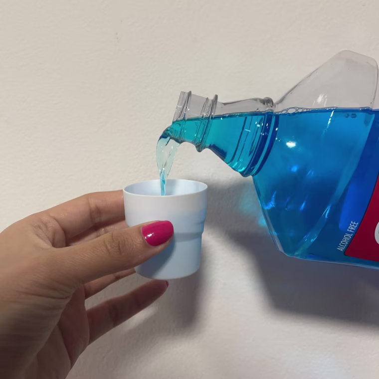 A woman’s hand holding a small white cup and pouring Colgate’s blue Total Active Prevention Mouthwash into it in front of a white wall.