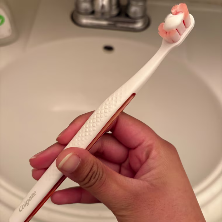 A woman’s hand holding a white and orange Colgate Total Active Prevention Foaming Clean Toothbrush with toothpaste on it over a sink.