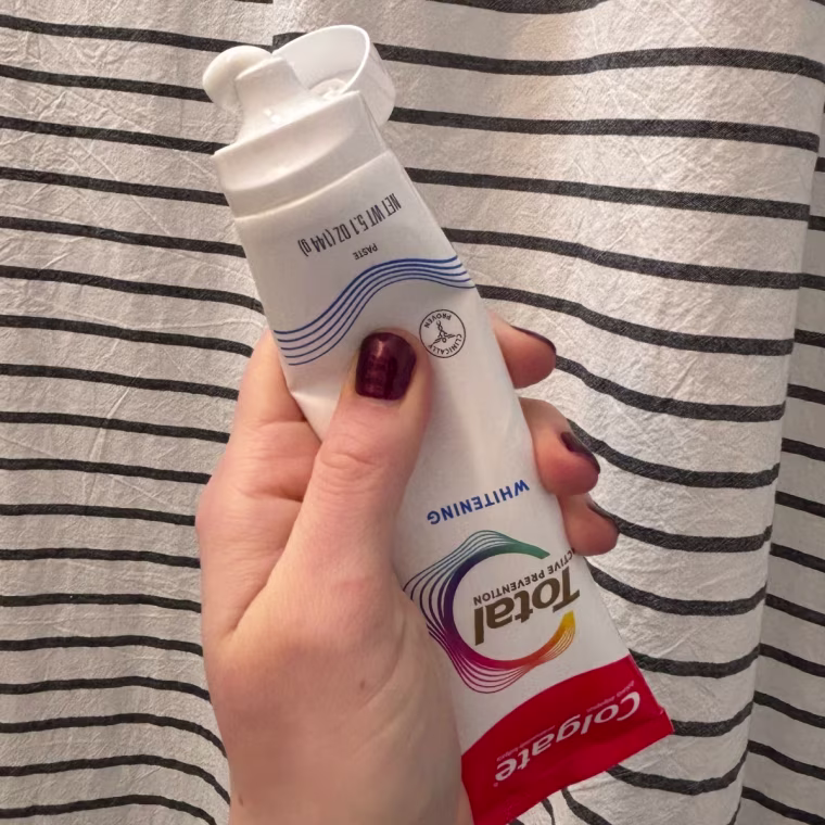 A woman’s hand squeezing Colgate Total Active Prevention Whitening Toothpaste out of a tube in front of a striped shower curtain.