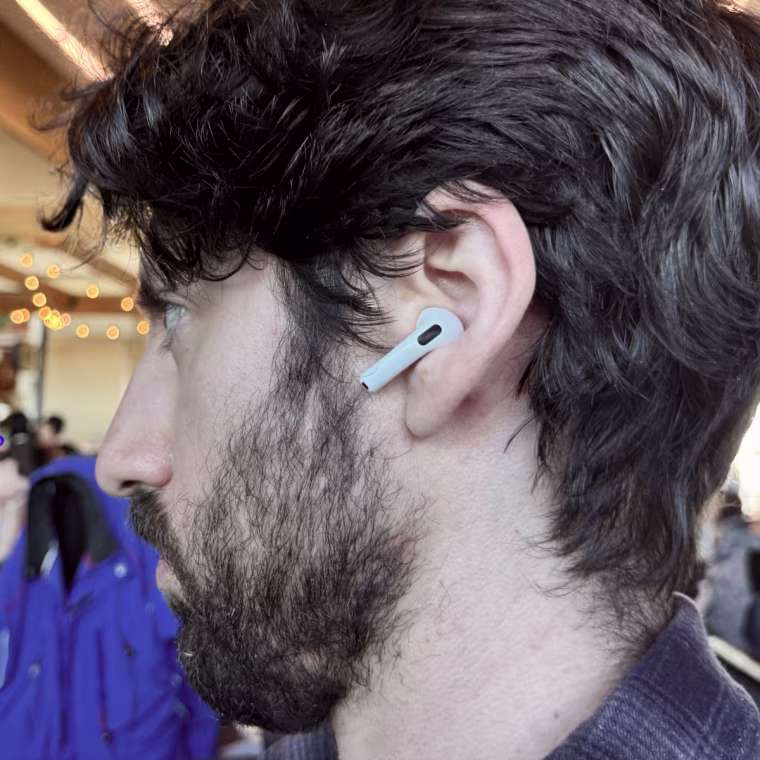 A side profile of the writer wearing Apple AirPods 4 with a crowded airport sitting area in the background.
