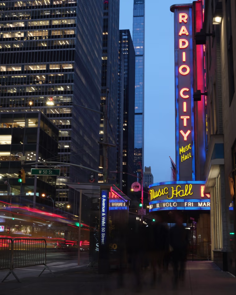 A wide angle photo of the Radio City Music Hall neon sign at night, with people and cars blurred in motion.