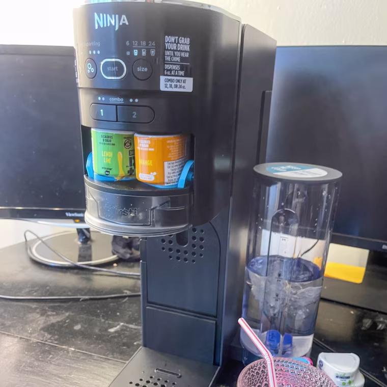 Ninja Thirsti Drink System Soda Maker sitting on a countertop beside a glass with a straw in it.