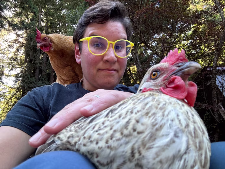 Christin New holds a chicken while another one stands on her shoulder, she wears yellow rimmed glasses