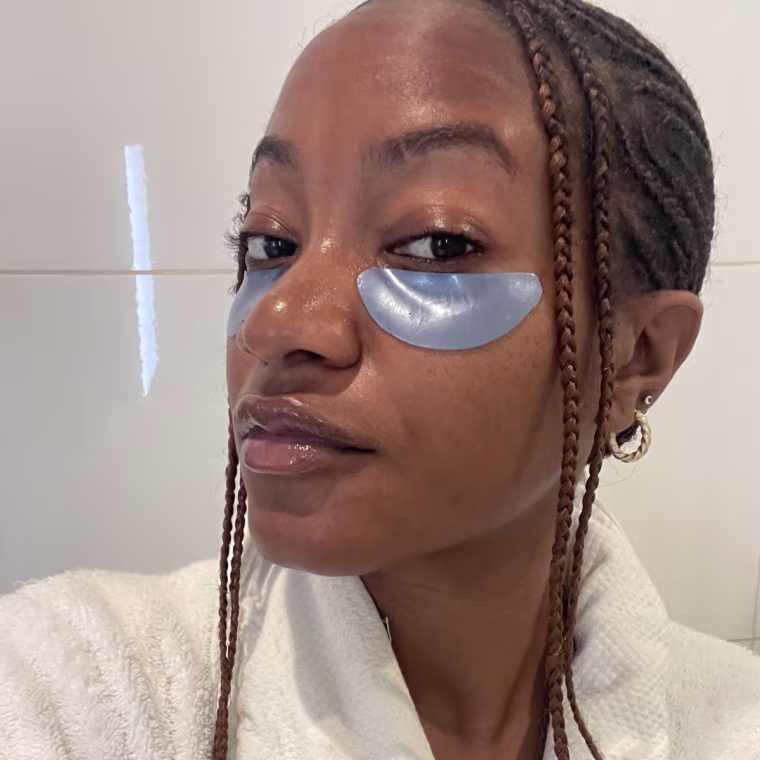Ashley Morris wearing the blue Summer Fridays Jet Lag Eye Patches, in a white robe against a white, tiled bathroom wall.