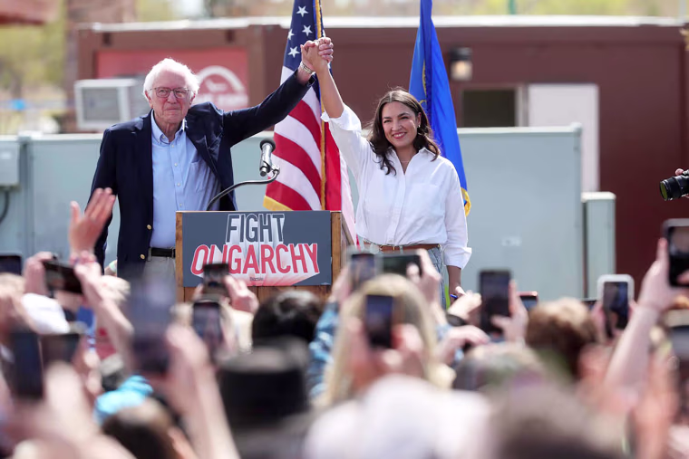 Bernie Sanders, left, and Alexandra Ocasio-Cortez hold hands and raise them to an audience onstage