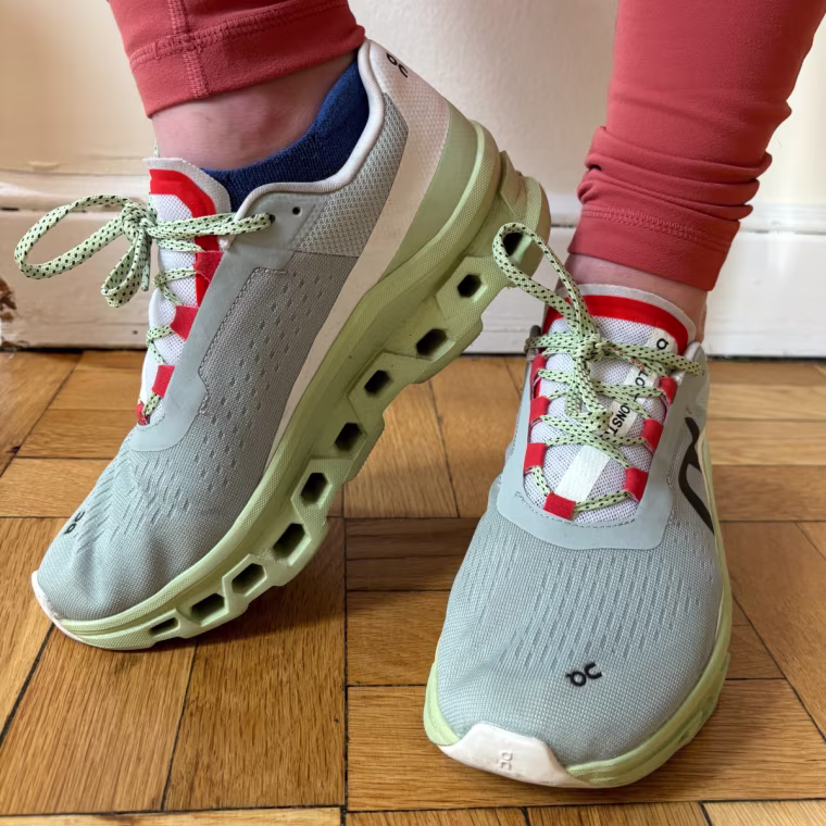A woman wearing On Cloudmonster sneakers on a wood floor in front of a white wall.