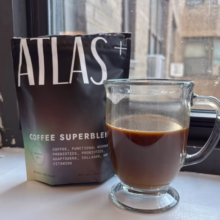 A bag of Altas+ Coffee Superblend next to a clear mug with a brown beverage inside it in front of a window.