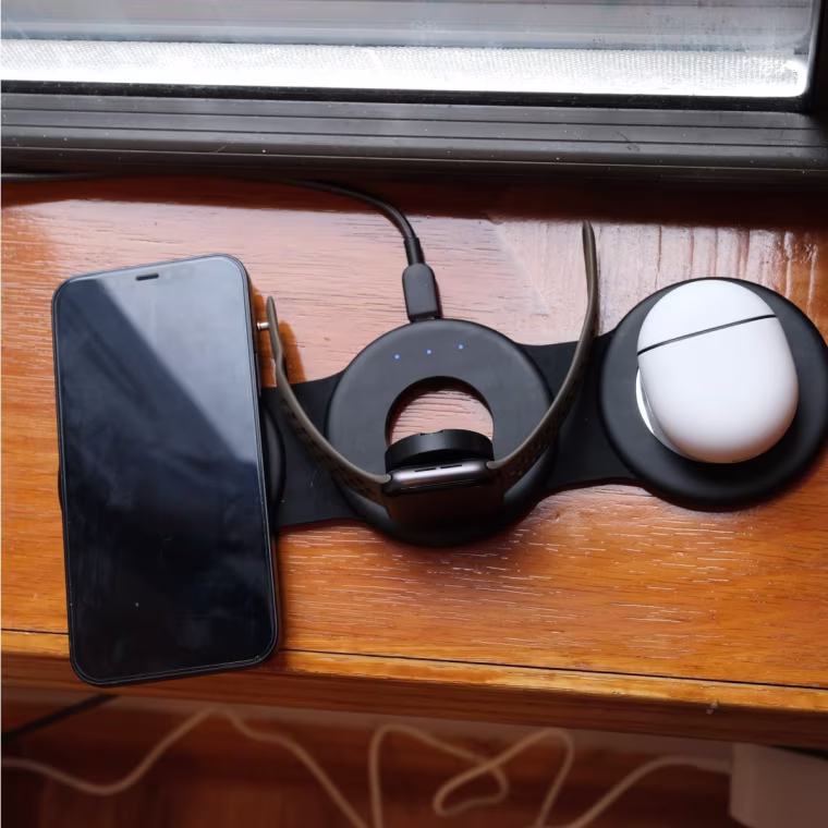 An iPhone, Apple Watch and earbuds case sitting on the Hicober charger on a windowsill.