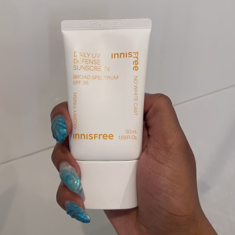 Innisfree Daily UV Defense Sunscreen being held by a hand with blue nails in front of a white tile wall.