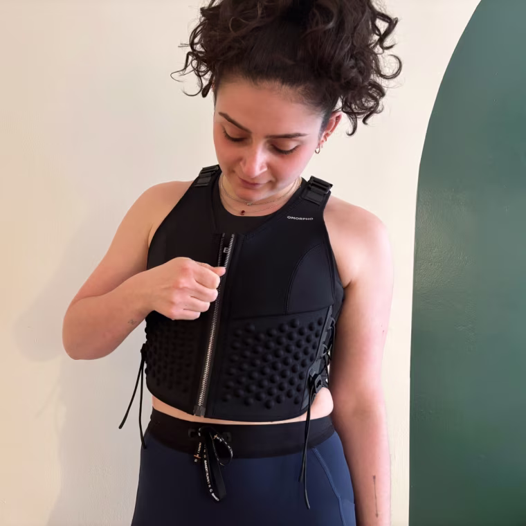 A woman wearing and zipping up Omorpho’s black G-Vest while standing in front of a white and green wall.