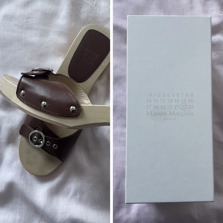Left: a white Maison Margiela shoe box on a light pink bedspread; Right: a hand holding one foot of a pair of Maison Margiela sandals with brown leather, silver studs and a light brown wood block heel, with the other foot on the pink bedspread behind it.