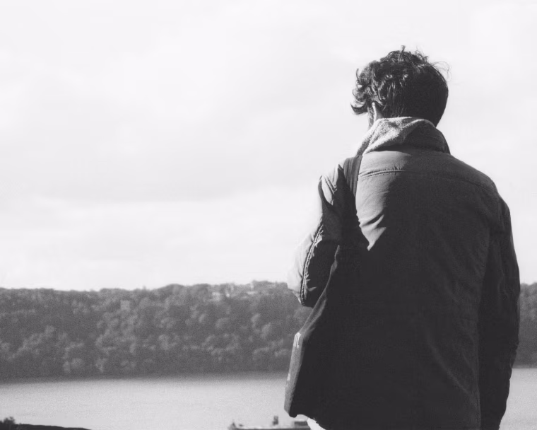 A black-and-white photo of a man facing away from the camera, looking at a river from on top of a cliffside.