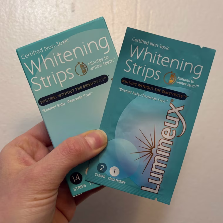 A person’s hand holding a box and package of Lumineux Teeth Whitening Strips.