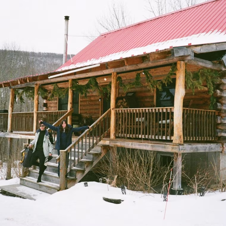 A photo of a large log cabin covered in snow. Two people are walking down the patio stairs.
