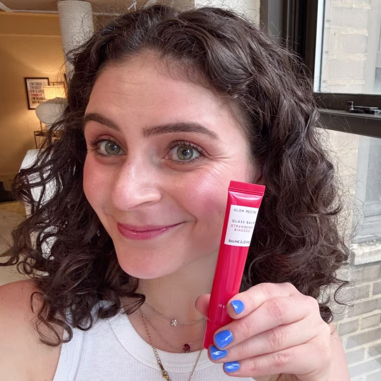 A woman holding a tube of Glow Recipe’s Glass Balm next to her face while wearing the balm on her lips.