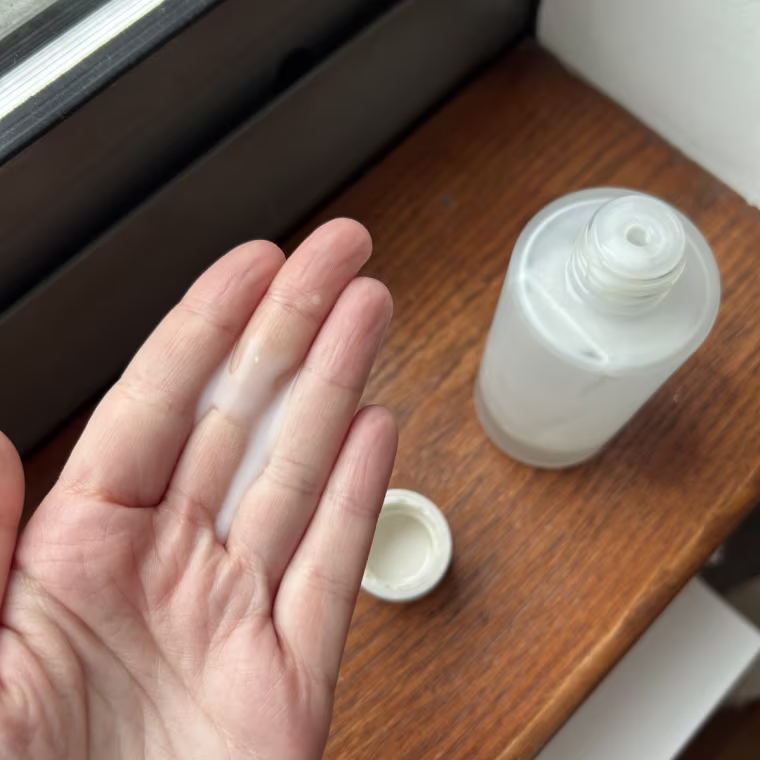 A hand holding a small pump of Ilia The Base Face Milk in beside the Ilia bottle on a windowsill.
