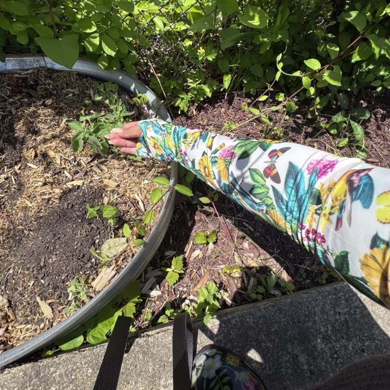Brown wearing the floral Uncommon Goods Protective Sleeve on her arm, held in front of a garden patch with soil, plants, and a fence in the back.