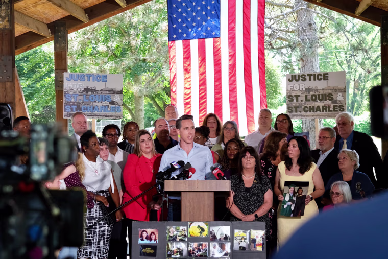Josh Hawley speaks at a podium outside, people surround him, and American flag hangs behind him