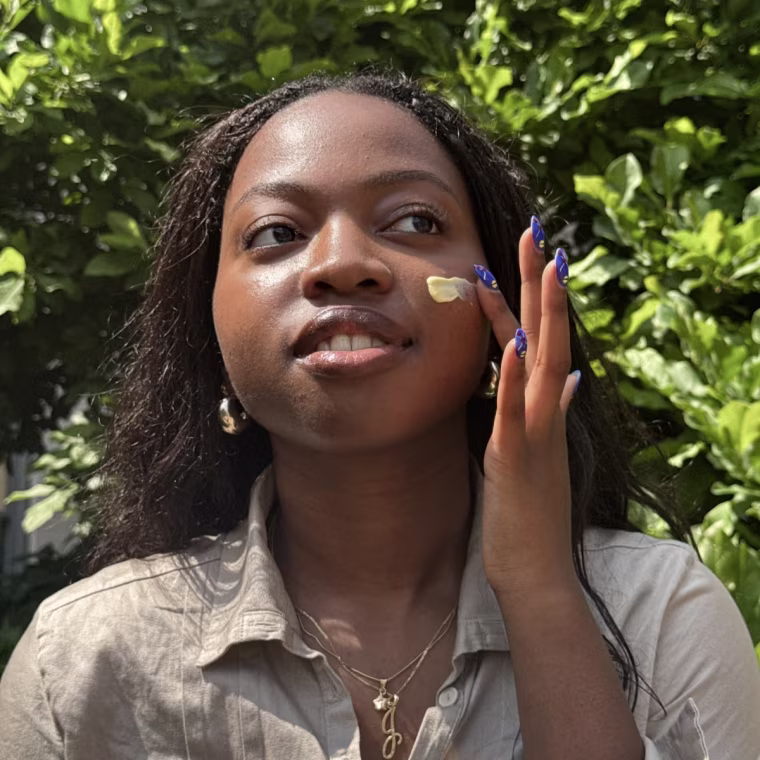 Alabi in front of a background of greenery, rubbing the Good Weather Skin Sun Cream into her face.