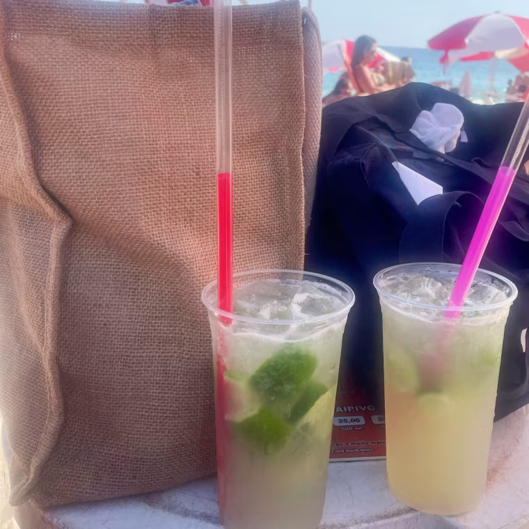 The Muji Jute bag on a white table beside a black tote bag, with two lime drinks in front and a beach in the background.