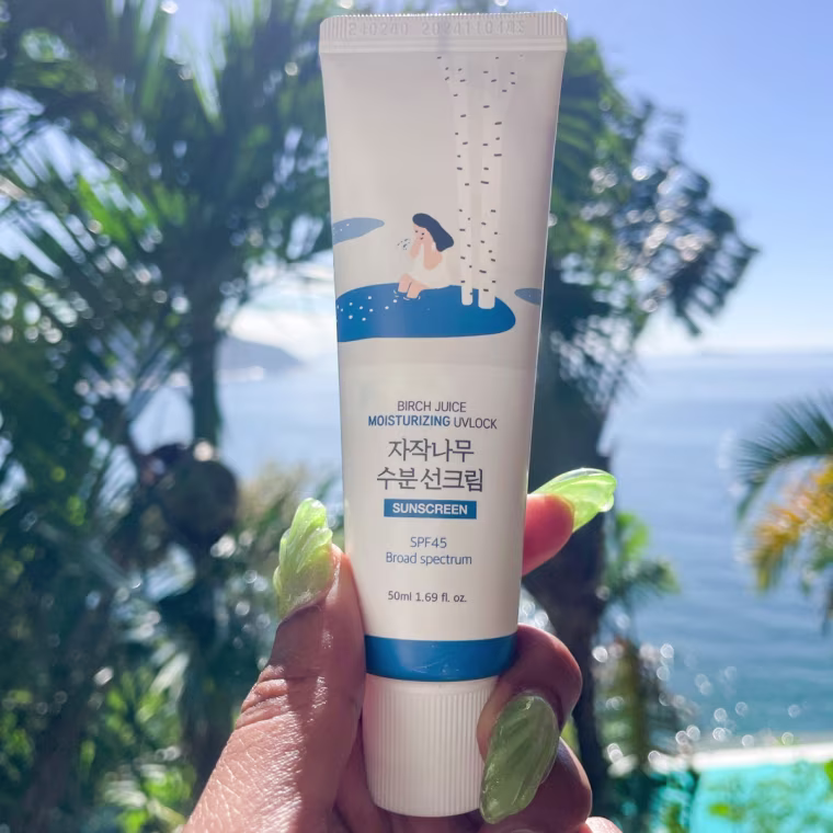A left hand with green nails holding the Round Lab sunscreen in front of an ocean background.