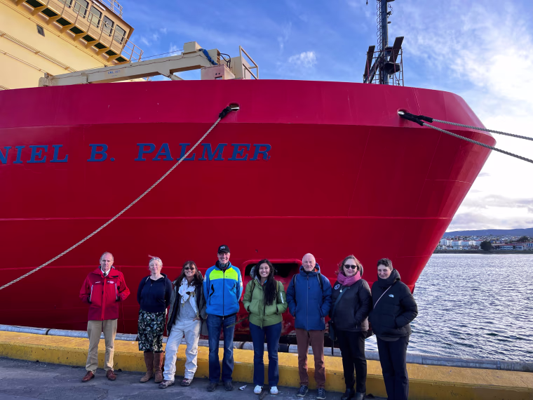 A row of eight people stand in front of a large red ship