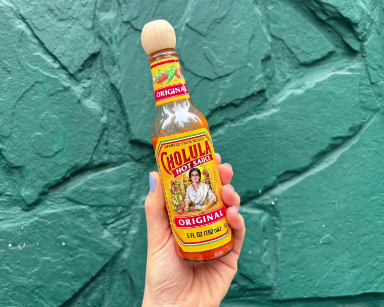 A person’s hand holding a bottle of Cholula in front of a teal wall.