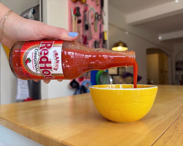 A hand pouring Frank’s Red Hot Original Hot Sauce into a small yellow bowl.