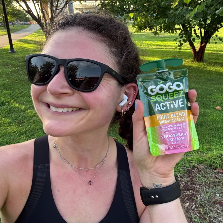 A woman holding a GoGo Squeez Active Fruit Blend Energy Release pouch outside next to her face.