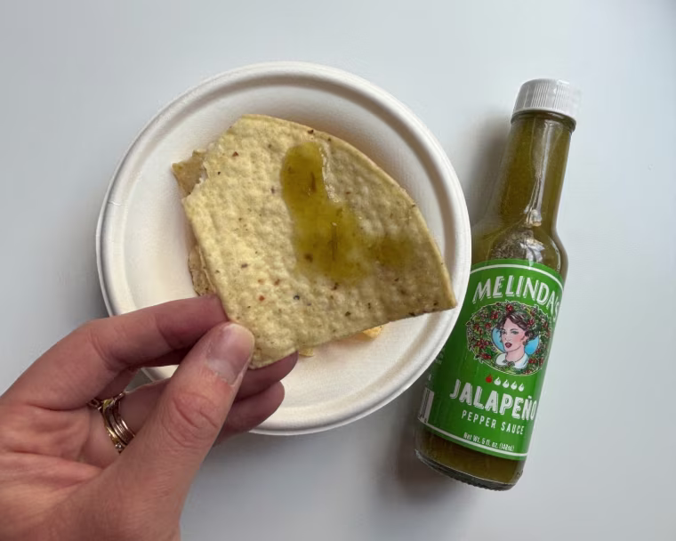 A hand holding a tortilla chip with Melinda’s Jalapeño Pepper Hot Sauce on it and a bottle of the sauce in the background.