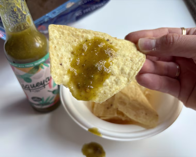 A hand holding a tortilla chip with Pisqueya’s Medium Heat Hot Sauce on it and a bottle of the sauce in the background.