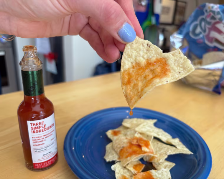 A hand shaking a few drops of Tabasco Original Red Pepper Hot Sauce onto a tortilla chip.